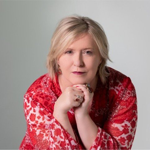Headshot of white woman with short blonde hair, wearing a red top and red jewellery leaning forward onto her hands